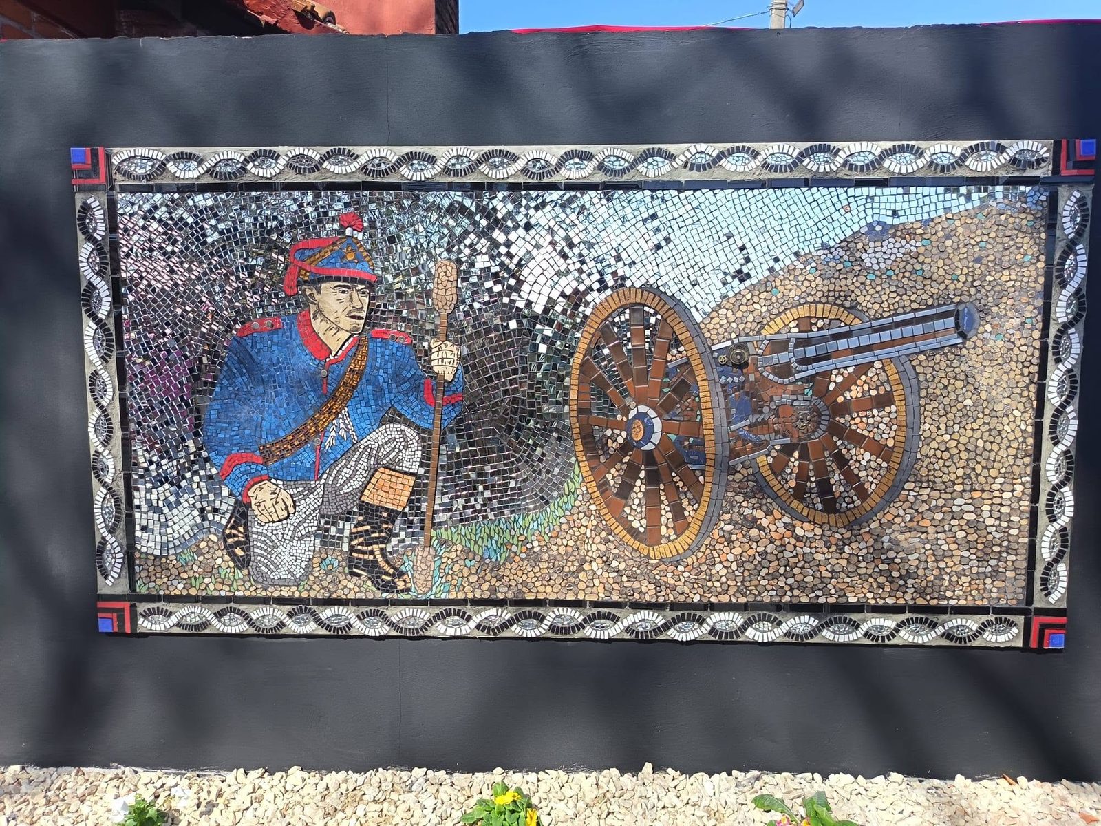 El mural: un artillero de uniforme histórico junto a un cañón, en teselas de cerámica, piedra y espejo, con una cenefa de espejos alrededor