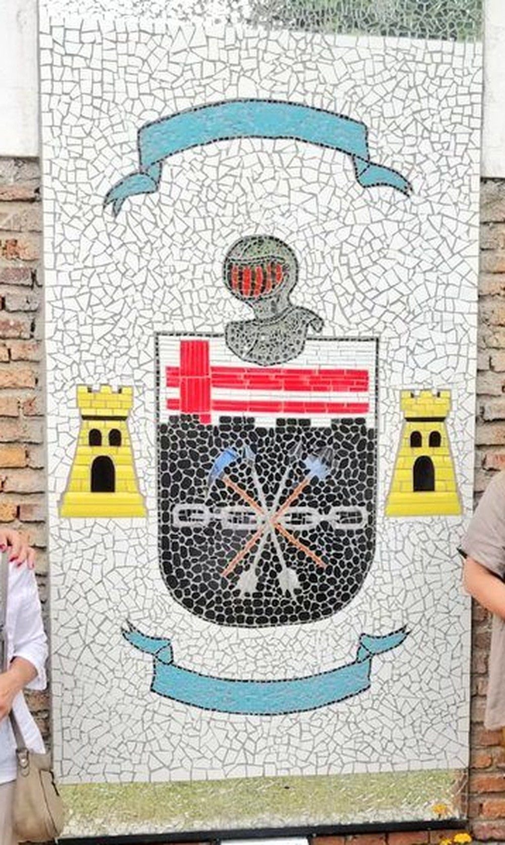 Detalle ampliado del escudo: yelmo con cimera, cintas azules, franjas rojas en ángulo, torres doradas almenadas, y picota y pala cruzadas sobre la cadena de siete eslabones