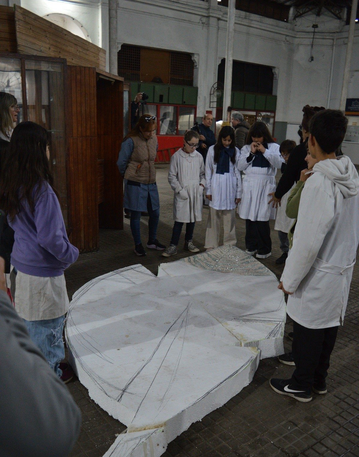 Base de la pluma vista desde arriba: forma recortada en telgopor, líneas de diseño a lápiz y un sector ya revestido con teselas espejadas; niños y adultos observando alrededor