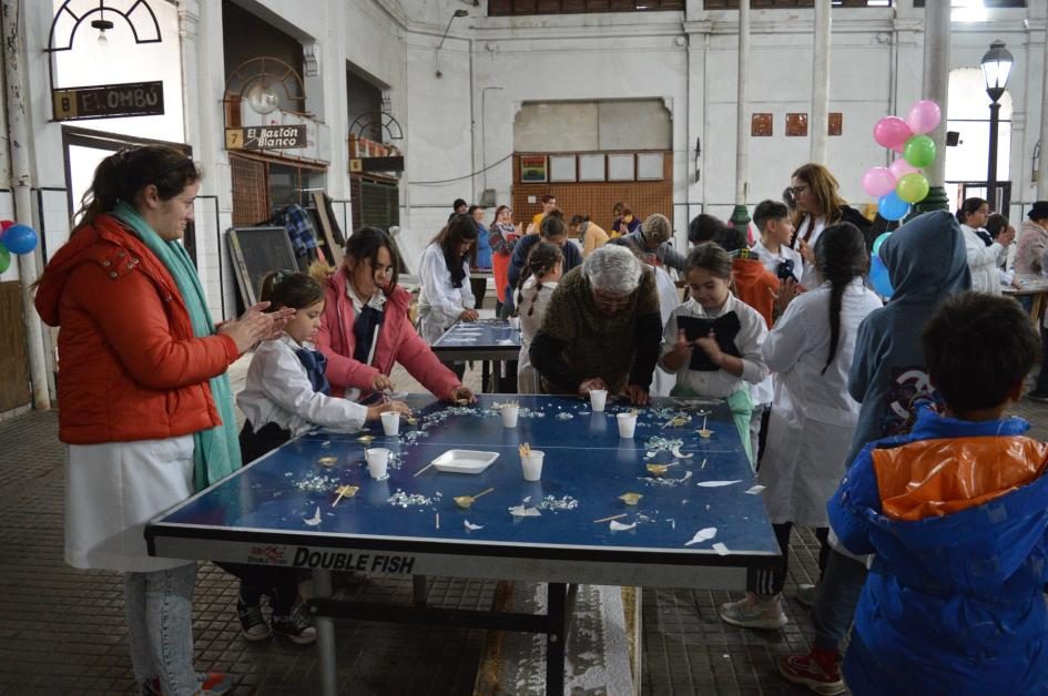 Otra mesa de trabajo con escolares y adultos seleccionando y pegando teselas, ambiente festivo con globos