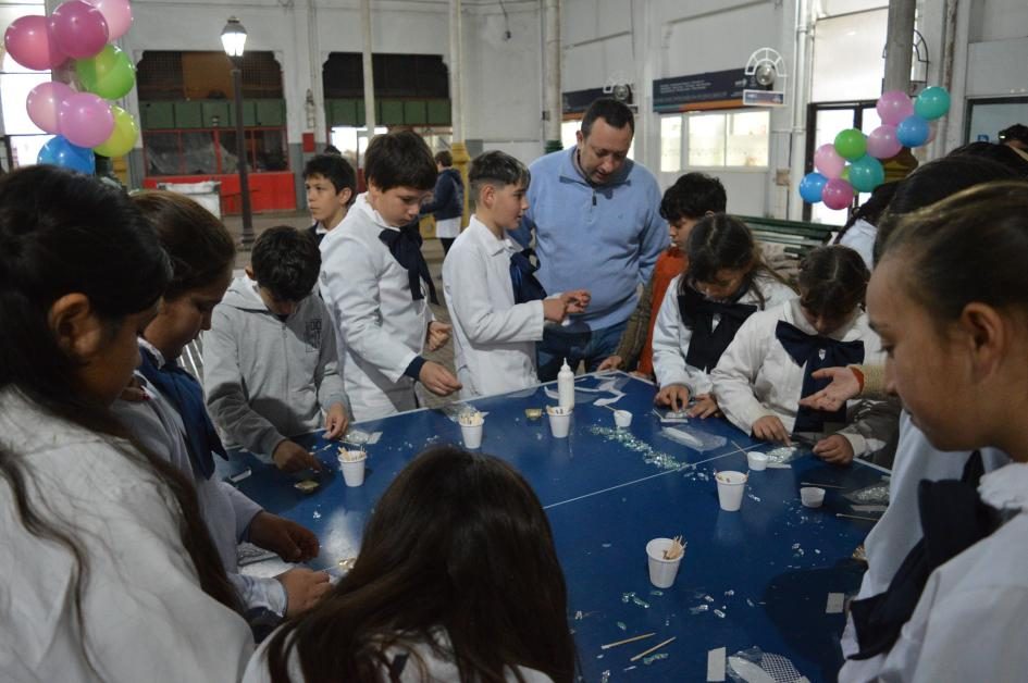 Escolares y un docente trabajando con teselas, pegamento y palillos sobre una mesa, con globos de decoración