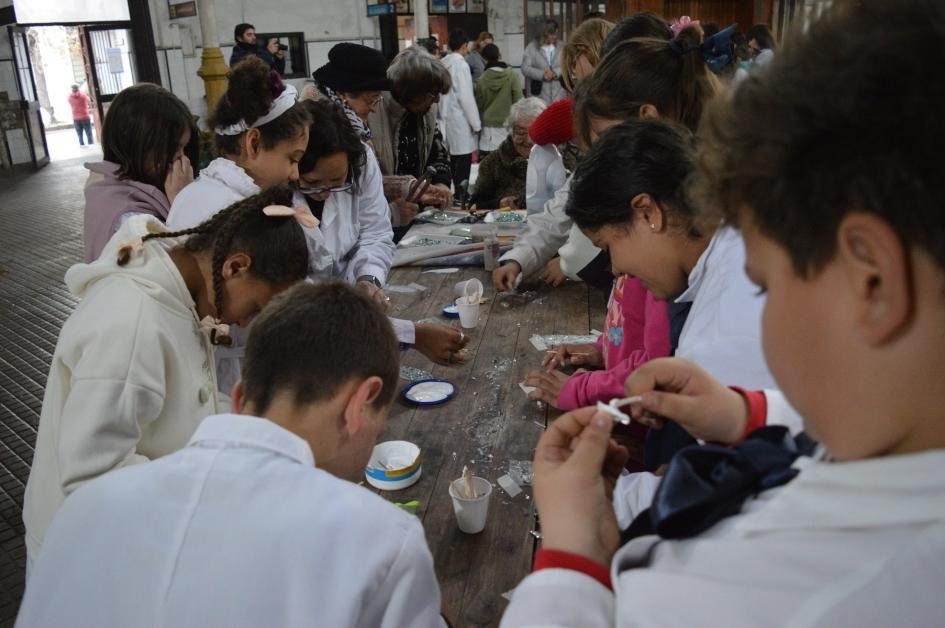 Escolares con guardapolvo blanco trabajando en una larga mesa de madera, pegando teselas