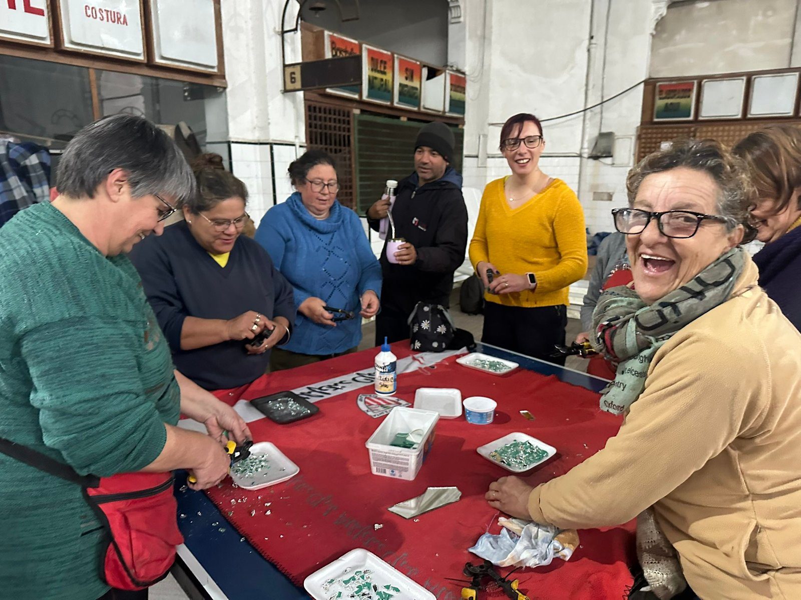 Integrantes del grupo, con mate, cortando y clasificando teselas de espejo sobre una mesa cubierta con tela roja, en el Mercado Municipal.