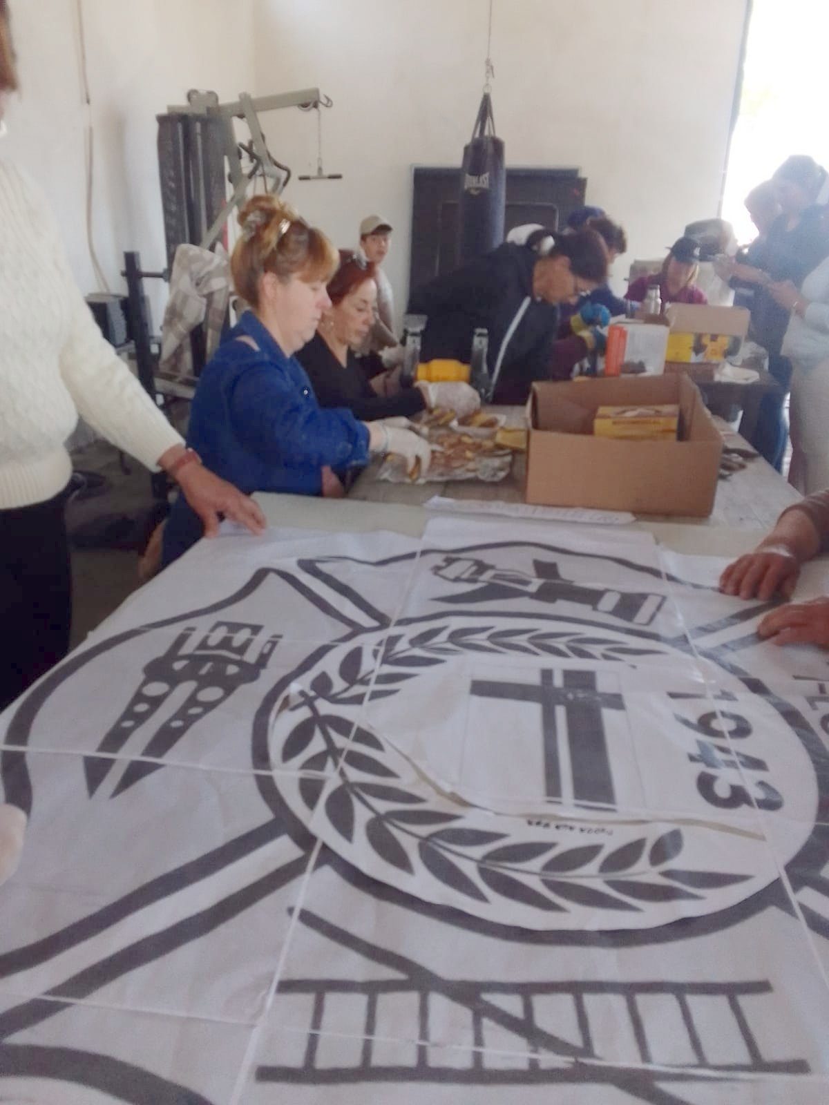 Plantilla de tela con el diseño del escudo calcado en negro (corona de ramas, cruz, escalera y el año "1943"), extendida sobre una mesa, con integrantes trabajando alrededor