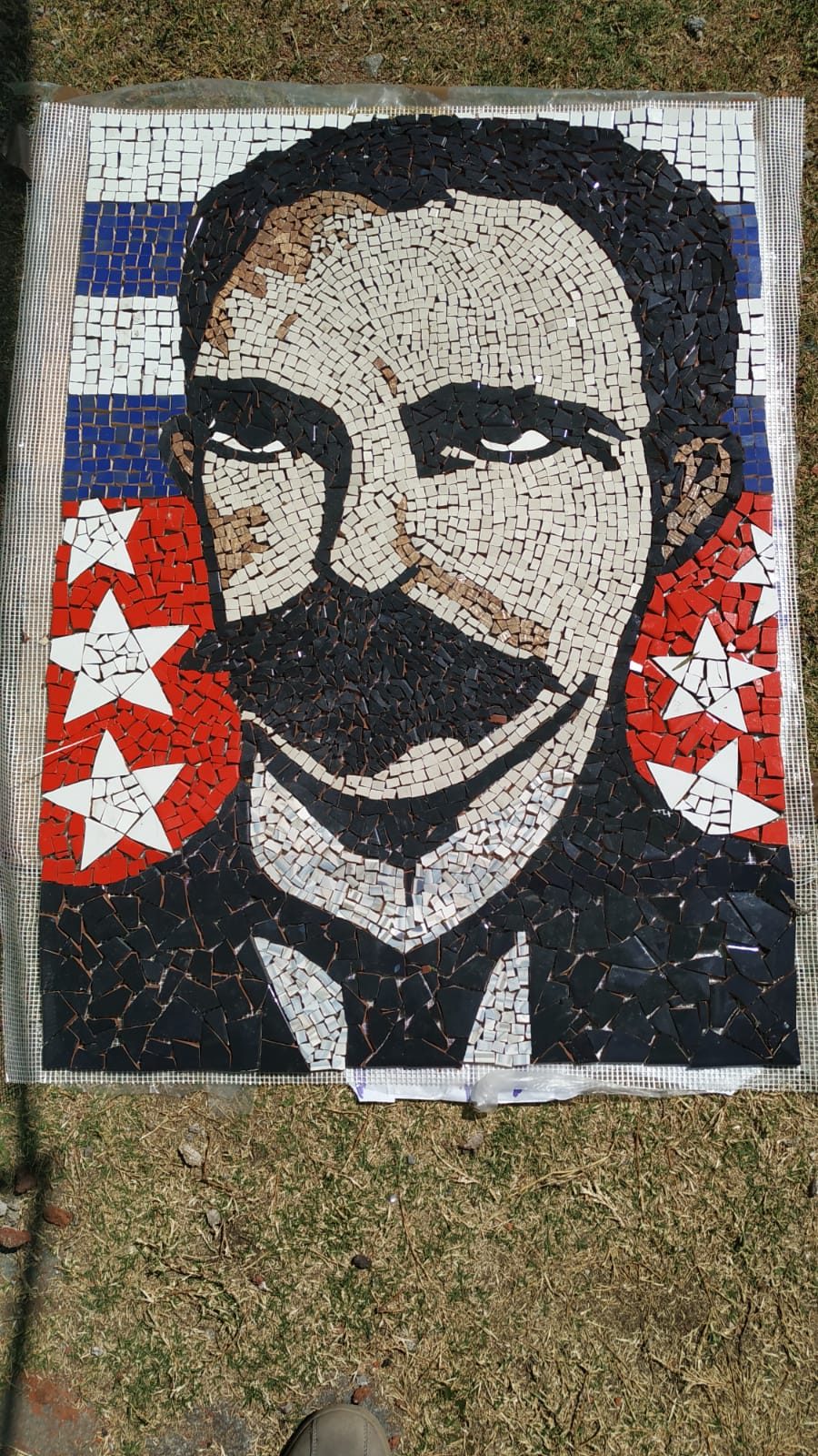 Mosaico figurativo del rostro de José Martí con fondo de bandera cubana (azul/blanco/rojo con estrellas), sobre malla, apoyado en el pasto