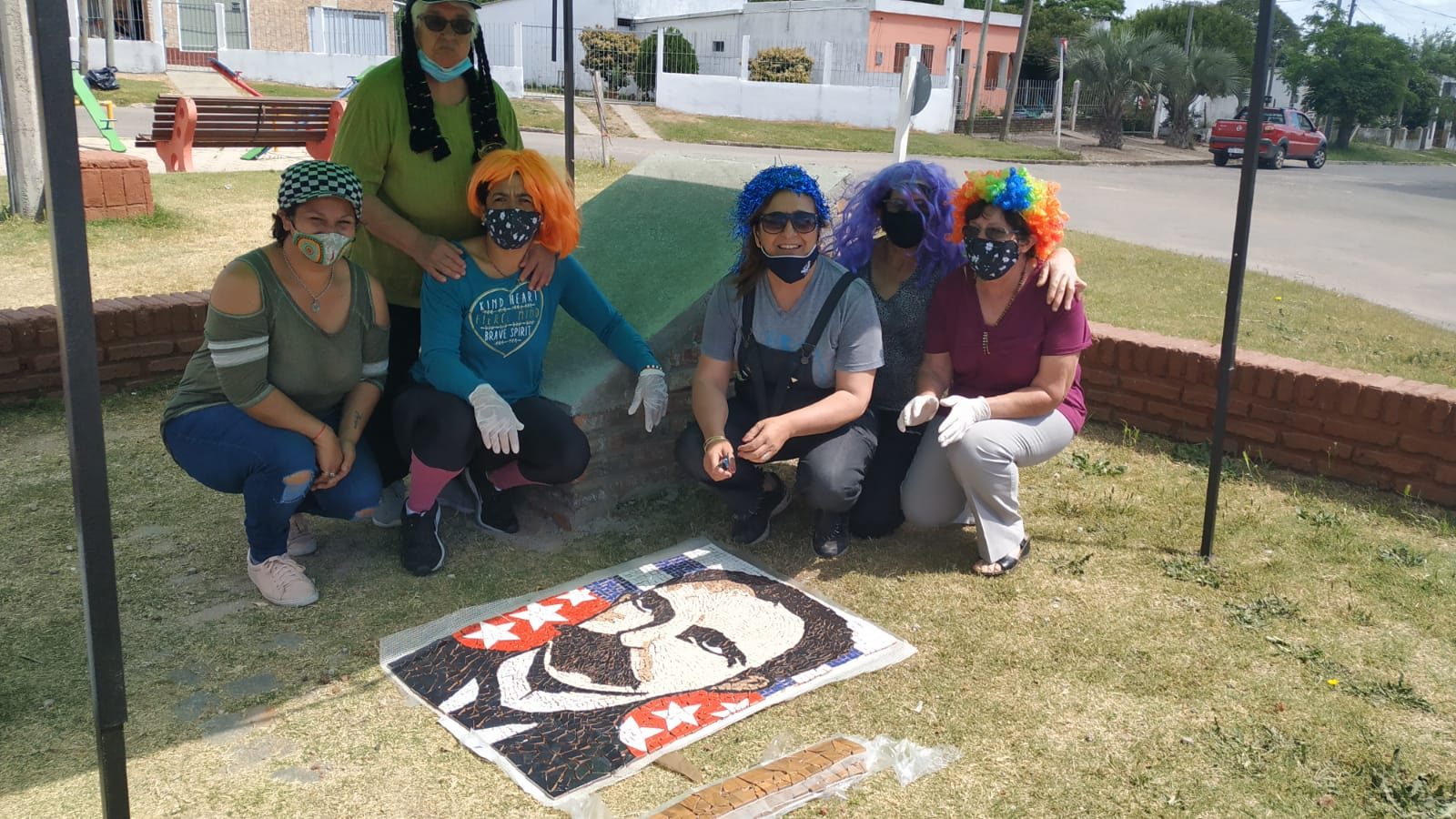 Seis integrantes de ReciclArte, con máscaras y pelucas de colores (contexto de pandemia/carnaval), en cuclillas junto al mosaico terminado del retrato de Martí, extendido sobre el pasto de una plaza con juegos infantiles al fondo
