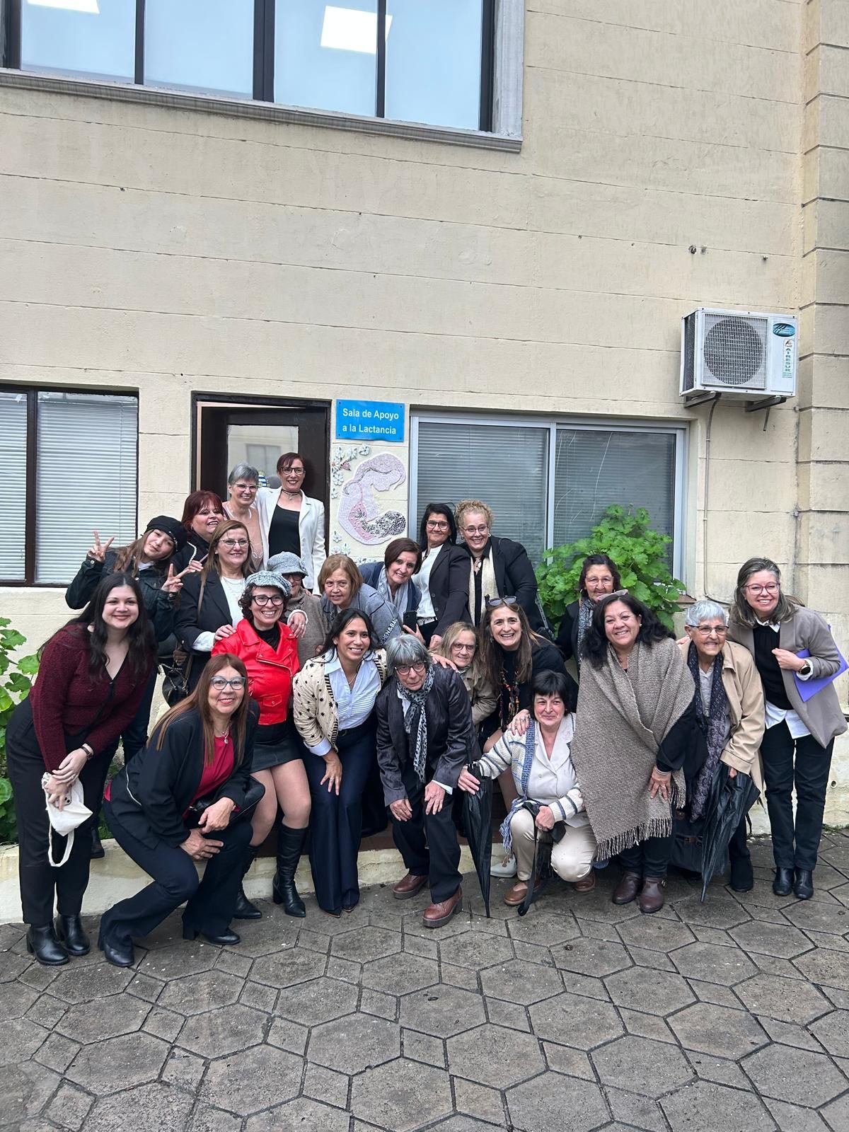 El grupo posa frente a la puerta de la sala de apoyo a la lactancia, con el mosaico de la madre y el bebé en la pared.