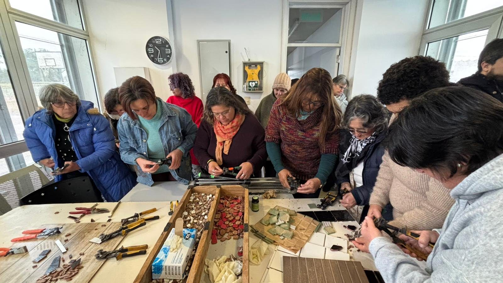 Mesa del taller con cajones de teselas de cerámica, azulejos partidos y tenazas; alrededor, integrantes del grupo cortando.