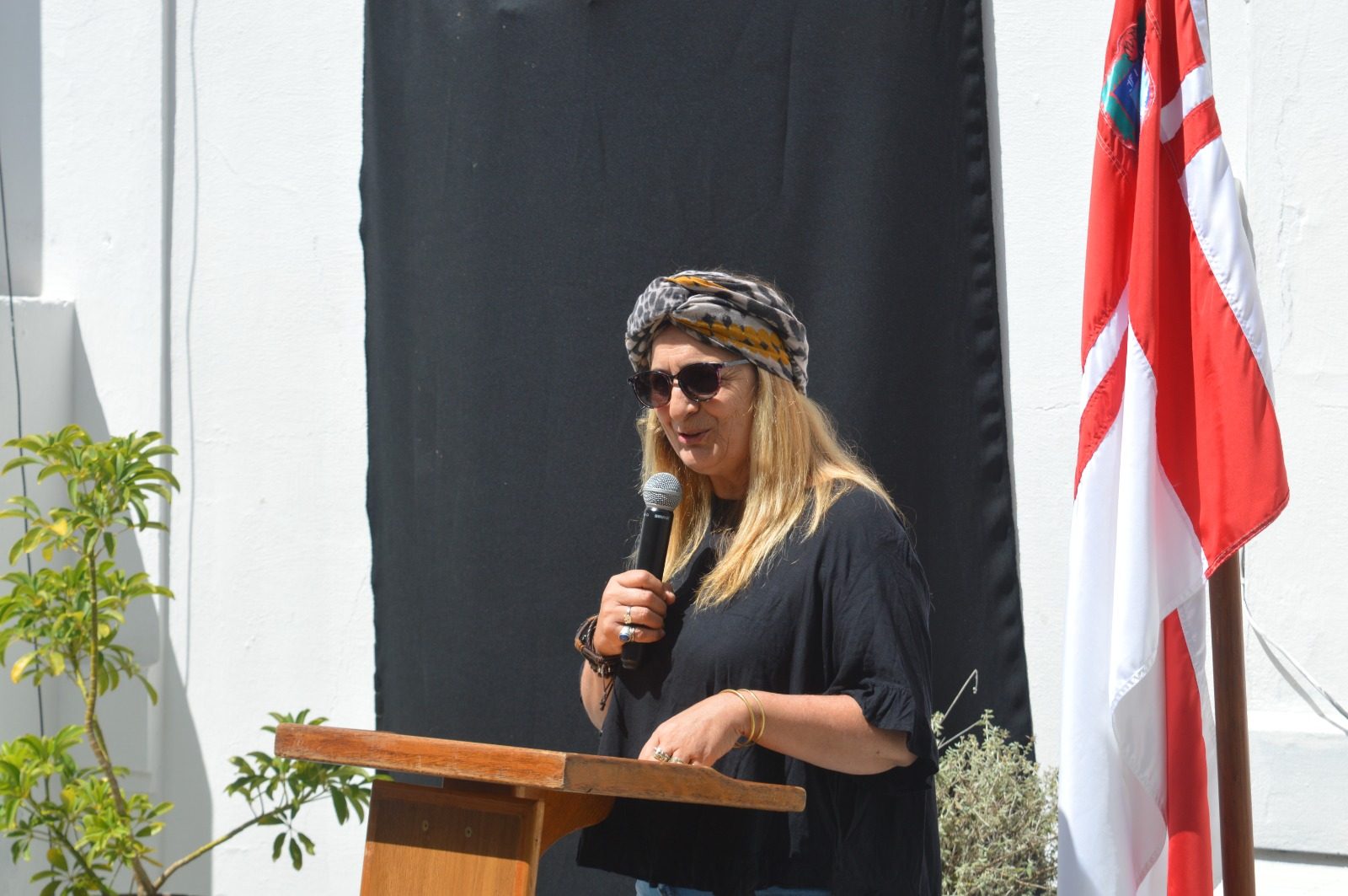 Alejandra Pérez (turbante estampado, anteojos de sol) hablando al micrófono en el atril durante el acto de inauguración