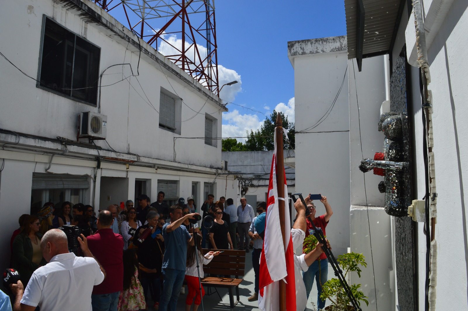 Plano general del patio del Edificio Comunal durante el acto: público, prensa (cámara de TVF) y el mural ya descubierto sobre la pared, banderas de Florida