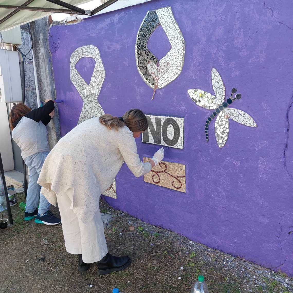 Tramo de pared pintada de violeta con símbolos en mosaico —una cinta/lazo blanco, una mano con una llama o corazón, una libélula y la palabra "NO"— mientras dos integrantes del grupo colocan y ajustan piezas