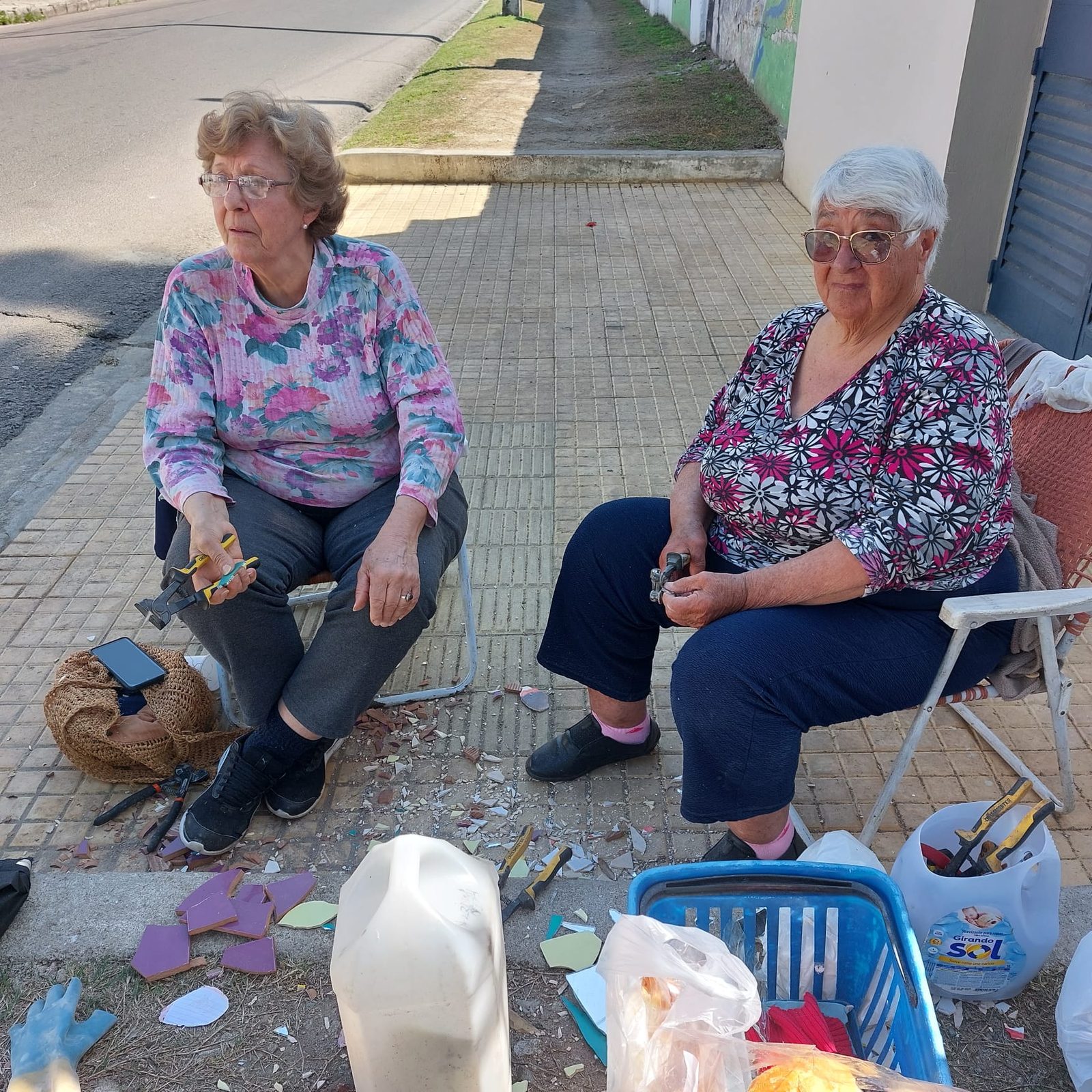 Dos integrantes de ReciclArte sentadas en la vereda, cortando y preparando piezas de cerámica con tenazas, con material y herramientas alrededor (foto de proceso, no muestra el mural en sí)