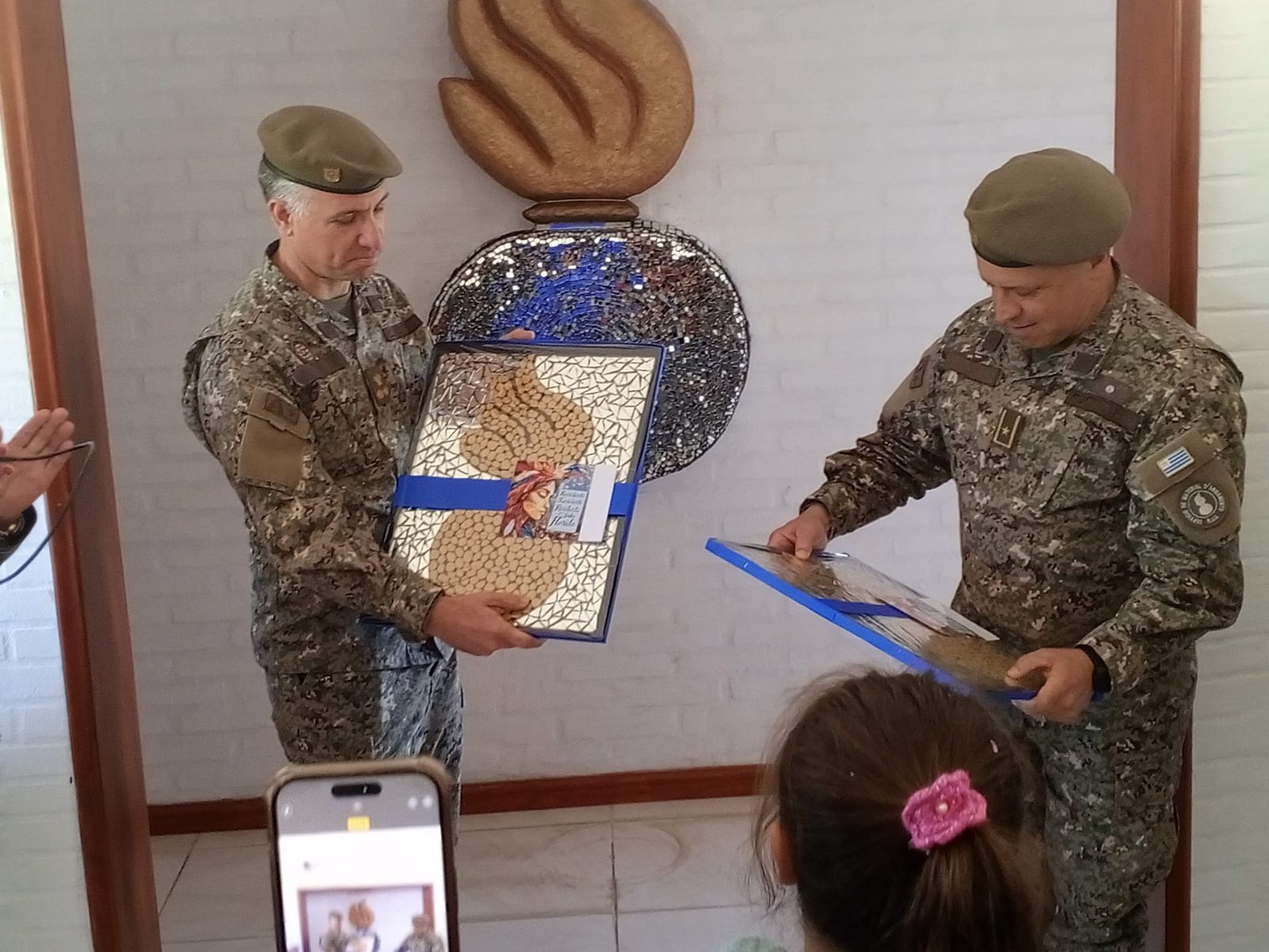 Dos oficiales del PESMA reciben un álbum/carpeta con tapa de mosaico como obsequio de ReciclArte, con la escultura de la llama de fondo