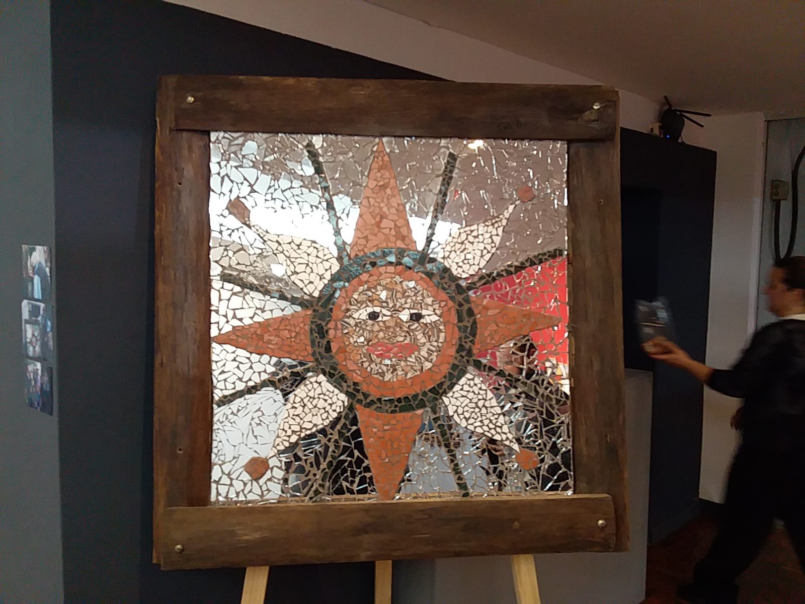 Vista frontal del mosaico circular ("sol" con rostro y rayos, fondo de espejo roto) montado sobre caballete de madera, dentro de la sala del MAVEA, con una persona pasando al fondo